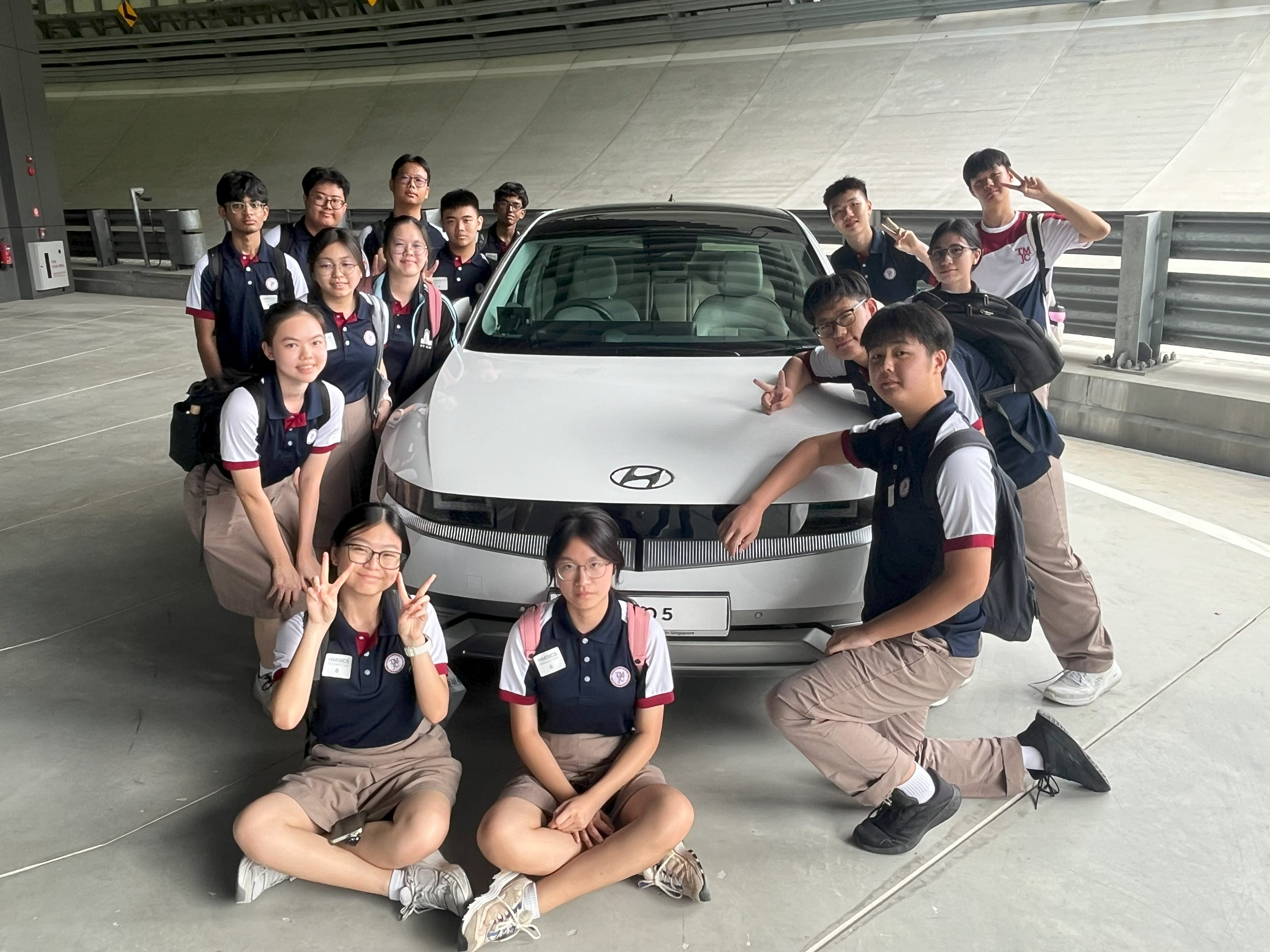 A group of about 15 students in school uniforms pose cheerfully around a white Hyundai electric car in an indoor carpark setting.
